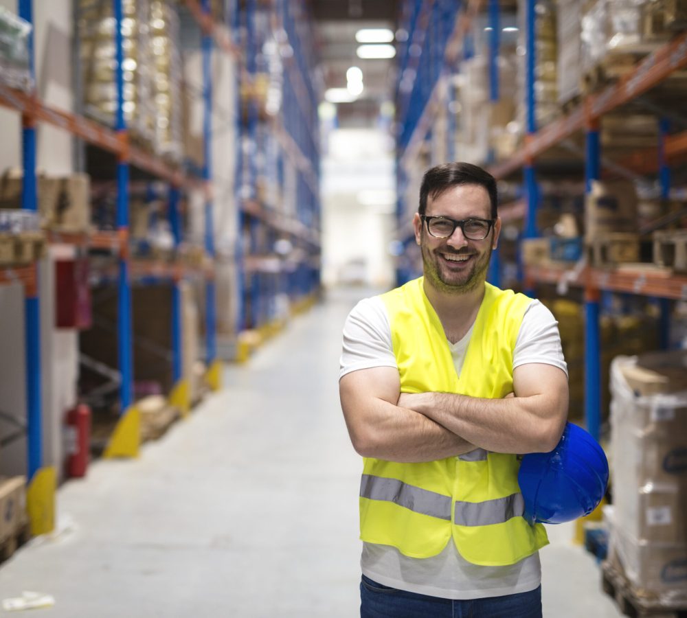 Portrait of middle aged warehouse worker standing in large warehouse distribution center with arms crossed.